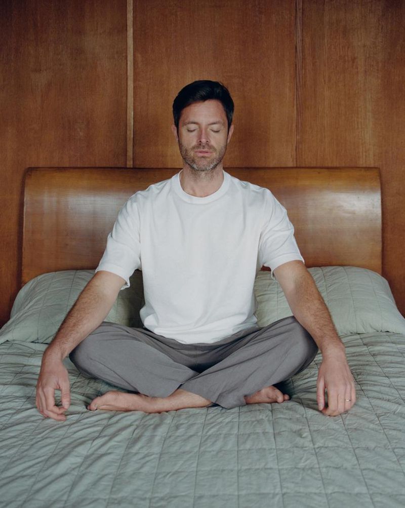 Man in a white t-shirt and gray pants sitting cross-legged on a bed in a meditative pose in front of a wood-paneled wall and headboard.