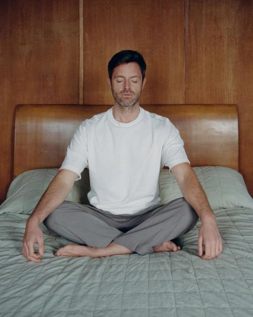 Man in a white t-shirt and gray pants sitting cross-legged on a bed in a meditative pose in front of a wood-paneled wall and headboard.
