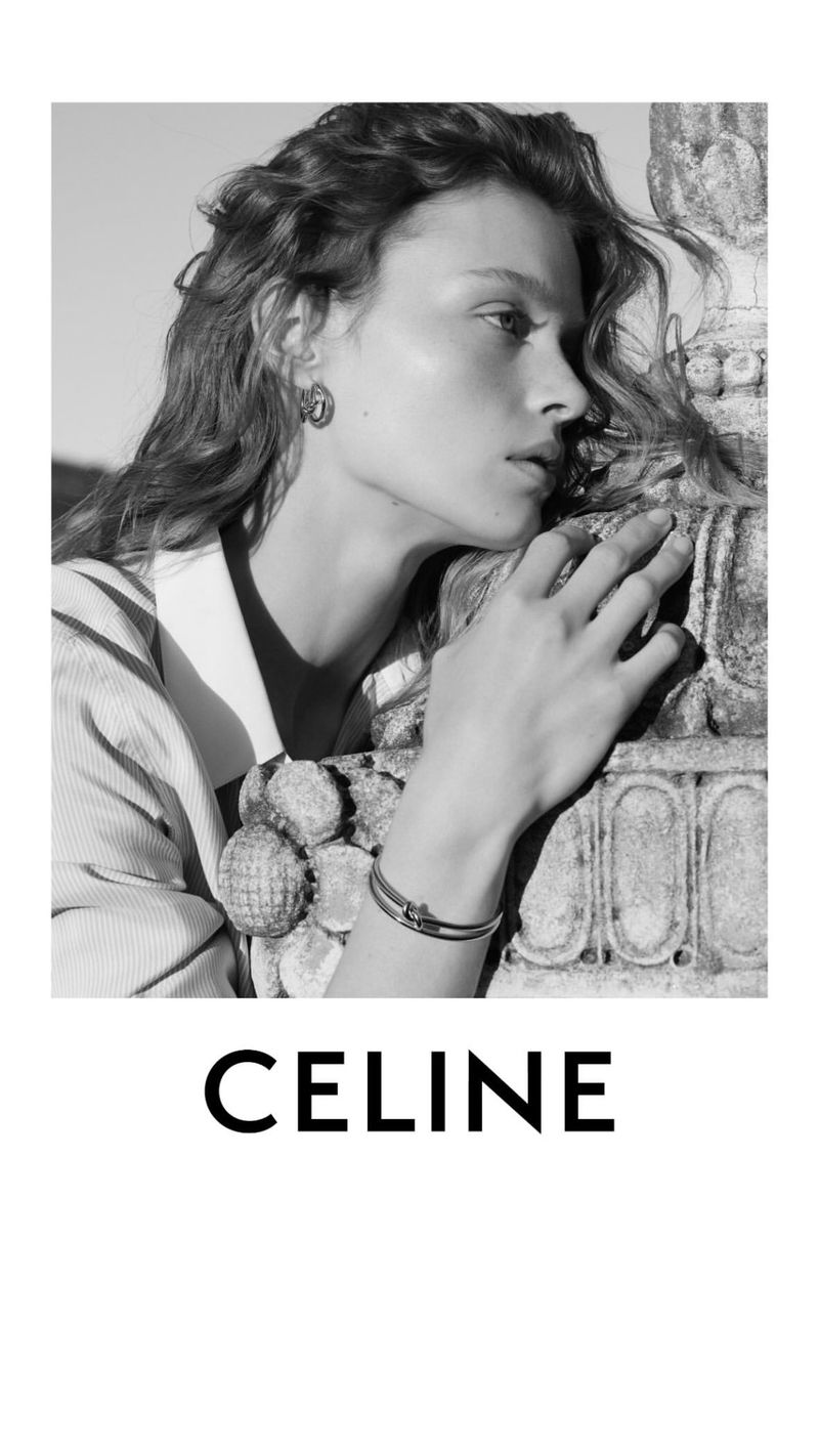 Black and white portrait of a woman wearing jewelry leaning against a decorative stone feature with the CELINE logo at the bottom.