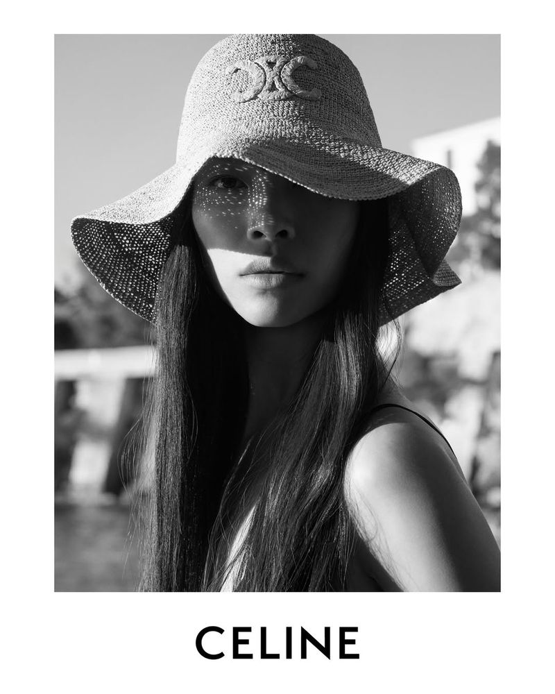 A woman poses in a black and white fashion shot, wearing a raffia hat with the Celine logo, against a blurred background on a sunny day.