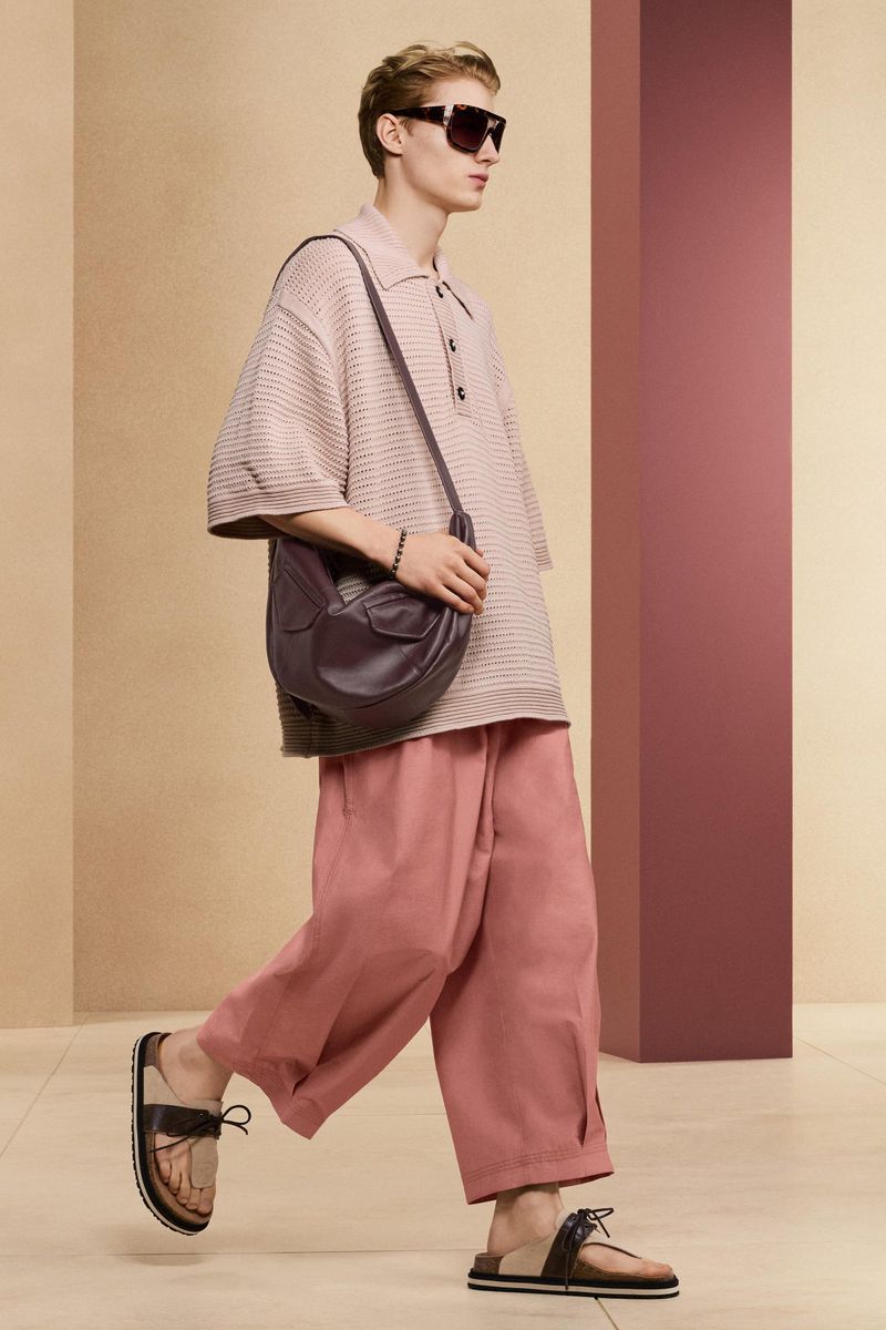 A fair-skinned male model poses in a studio wearing a light pink buttoned shirt, pink pants, brown sandals, sunglasses, and a large leather bag.