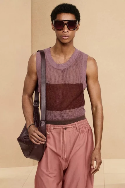 Man wearing a sleeveless knit shirt and sunglasses, posing indoors. He's carrying a leather bag in a fashion style look.