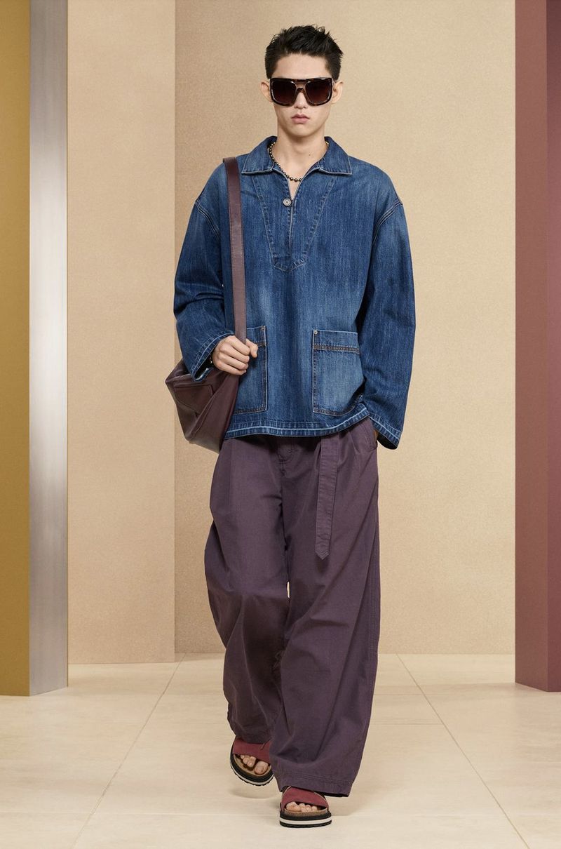Male fashion model in denim top, wide-leg pants, and sandals walks against a neutral studio backdrop with a messenger bag.