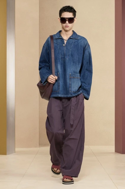 Male fashion model in denim top, wide-leg pants, and sandals walks against a neutral studio backdrop with a messenger bag.