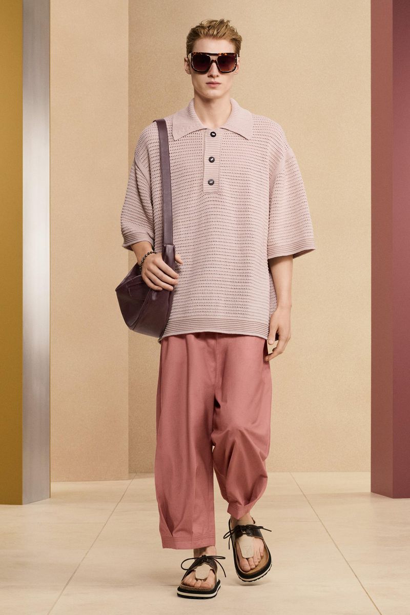 Male fashion model wearing a short-sleeve collared shirt, trousers, sandals, and shoulder bag, posing in a minimalist photo studio.