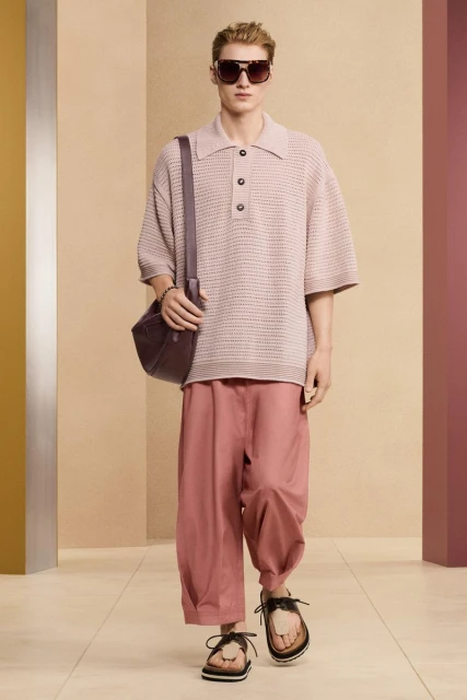 Male fashion model wearing a short-sleeve collared shirt, trousers, sandals, and shoulder bag, posing in a minimalist photo studio.