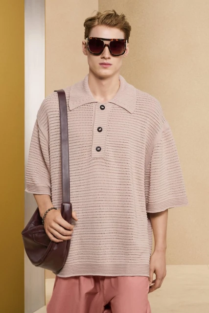 Male fashion model wearing a tan, knitted polo shirt, maroon slacks, and tortoise shell sunglasses while posing in a studio.