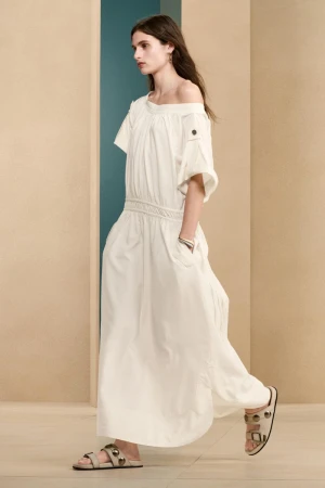 Model wearing an off-the-shoulder white dress and studded sandals against a beige architectural wall