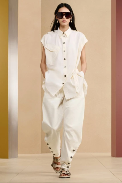 Runway fashion show featuring a female model in a sleeveless white button-down shirt, white pants, sunglasses, and sandals.
