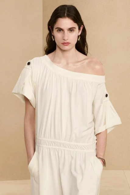 Fashion model poses in a cream-colored off-the-shoulder dress with short sleeves against a neutral backdrop. Studio shot, portrait.