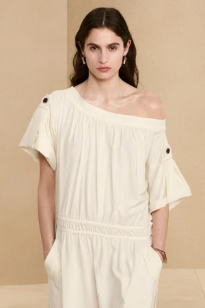 Fashion model poses in a cream-colored off-the-shoulder dress with short sleeves against a neutral backdrop. Studio shot, portrait.