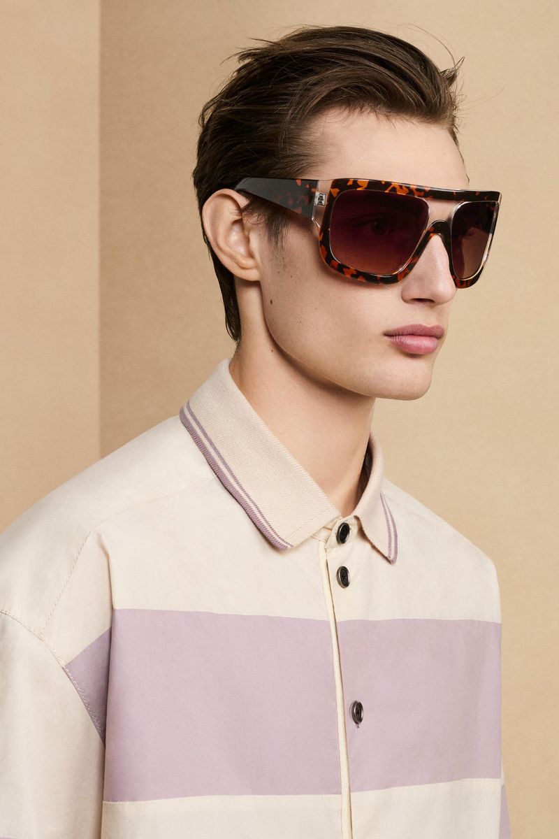 Close-up of a young adult man wearing large, square-framed, tortoise-shell sunglasses, sporting a fashionable retro shirt against a neutral backdrop.