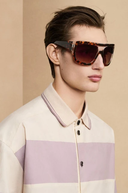 Close-up of a young adult man wearing large, square-framed, tortoise-shell sunglasses, sporting a fashionable retro shirt against a neutral backdrop.