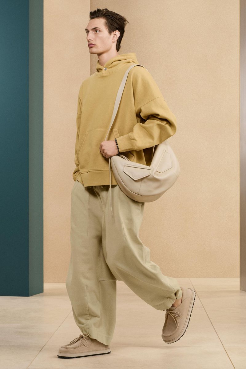 Full shot of a male model in a mustard hoodie, tan pants, and matching shoes, walking with a beige rounded bag on his shoulder.