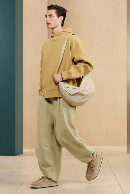 Full shot of a male model in a mustard hoodie, tan pants, and matching shoes, walking with a beige rounded bag on his shoulder.