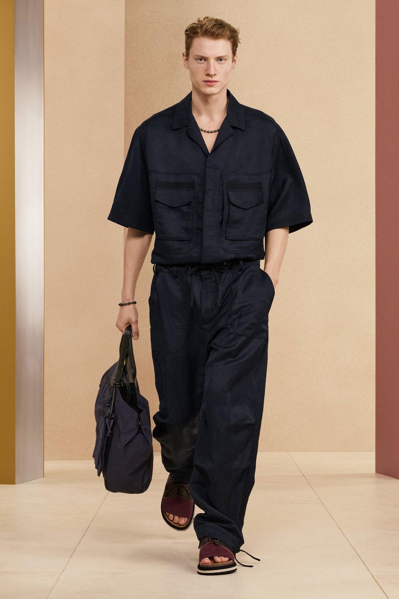 Full-length shot of a blond male model in an navy camp shirt and drawstring pants, walking on a light-colored floor holding a tote bag.