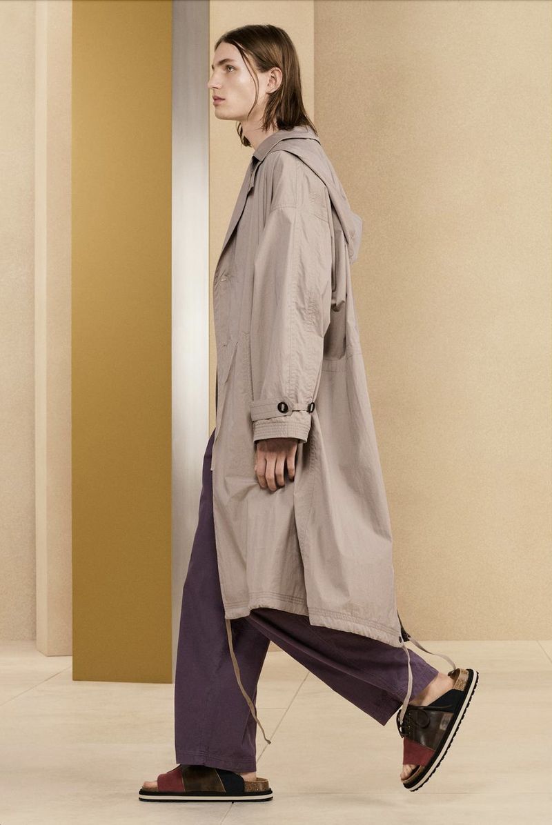 A person walks in a studio setting wearing a hooded tan coat, loose purple pants, and patchwork sandals against a two-tone backdrop.
