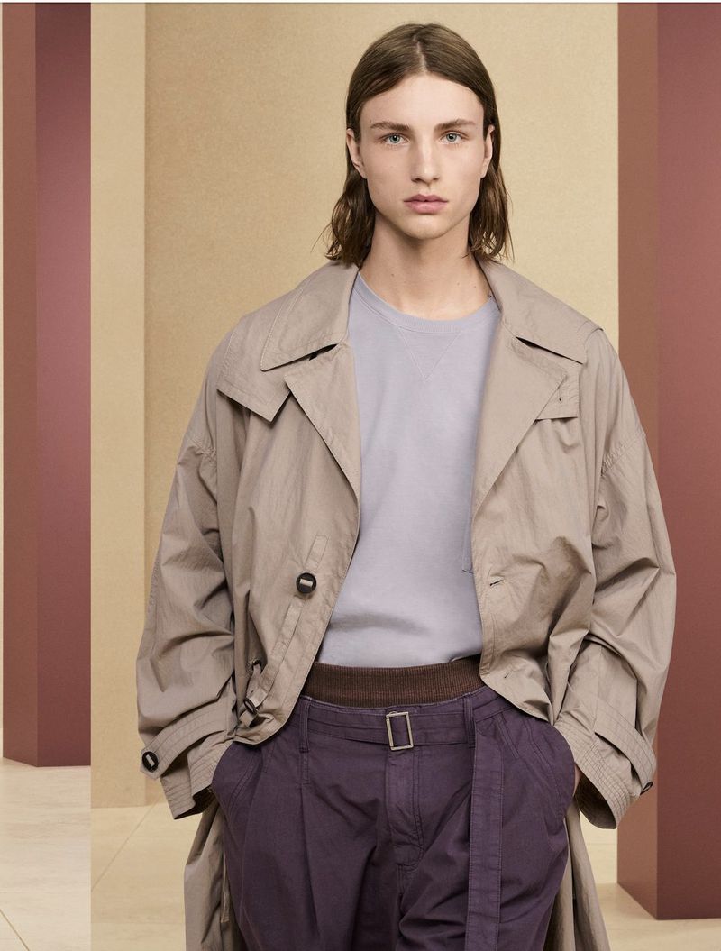 Portrait of a male fashion model wearing a trench coat and pleated pants against a background of beige and maroon panels.