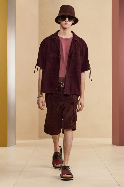 Male model in burgundy suede shirt and shorts with bucket hat on runway. Neutral color block background. Menswear fashion shoot.