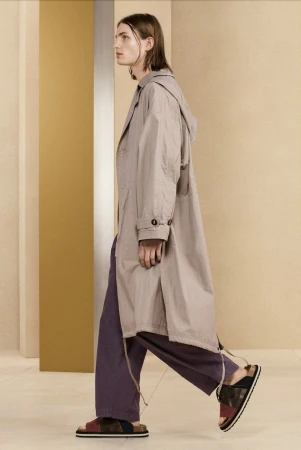 Profile view of a model walking in an oversized beige coat and purple trousers against a minimalist studio background.