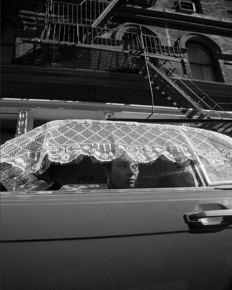 Black and white shot of a person in a car, lace draped on the windshield, set against a building with a fire escape.