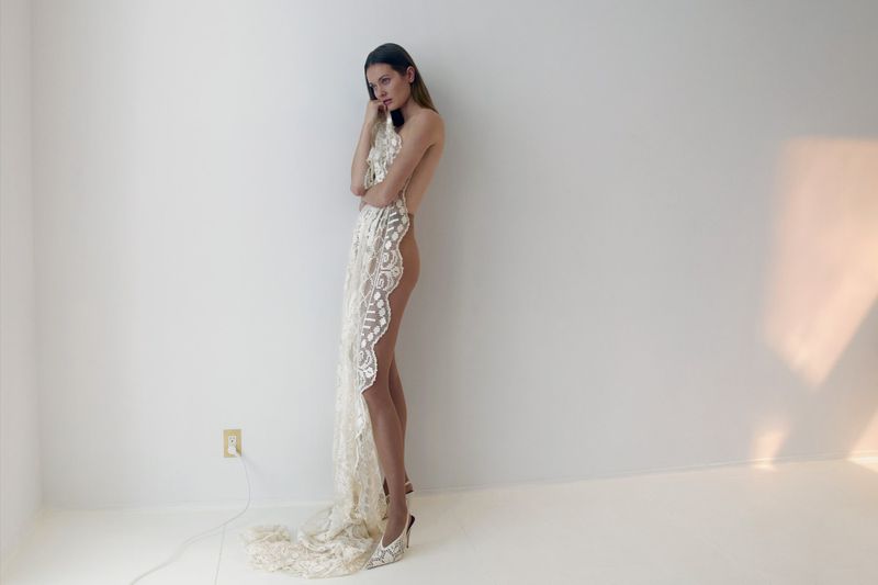 A woman in high heels draped with a lace fabric in a minimalist setting, illuminated by a soft, diffused light from an unseen source.