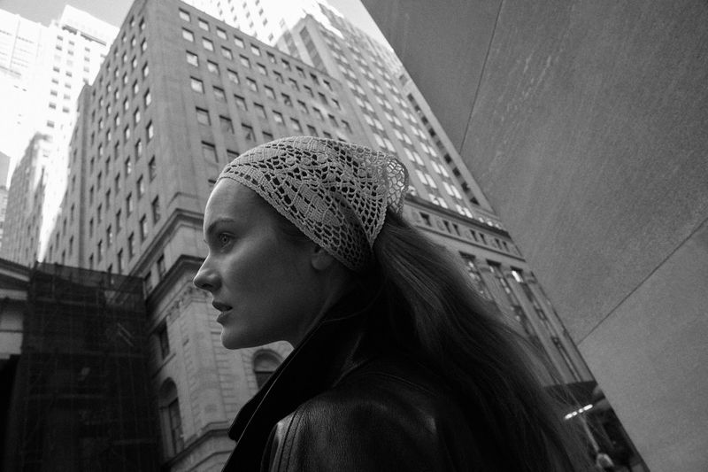 Black and white portrait of a woman with a headscarf, gazing to the left, with a tall building dominating the background.