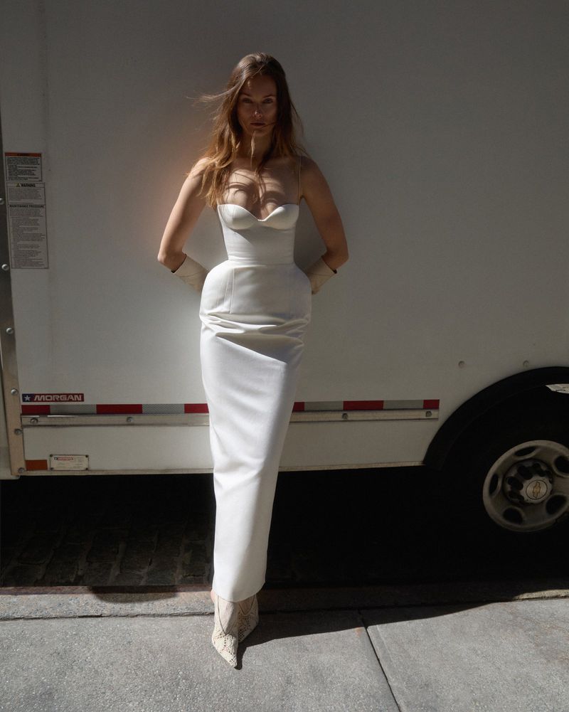A woman wearing a white dress and printed heeled boots poses next to a white trailer with concrete ground in an outdoor setting.