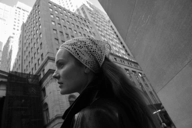 Model wearing a patterned headscarf and dark jacket outdoors against towering city skyscrapers.