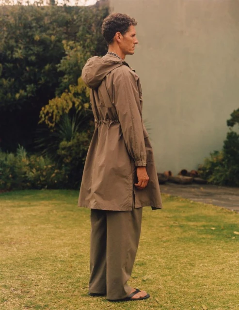 Adult male wearing a hooded khaki jacket and matching trousers, standing in a grassy outdoor setting with lush greenery in the background.