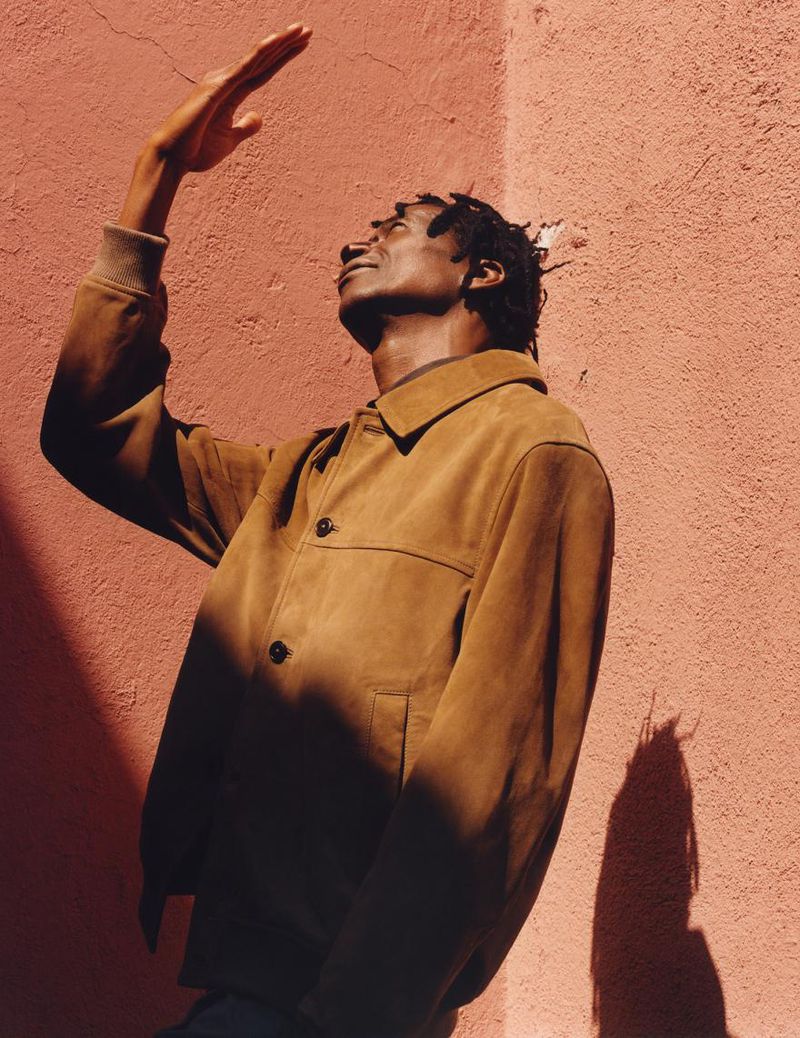 Portrait of a Black man in a brown jacket shielding his eyes with his hand in front of a salmon colored wall with a shadow.