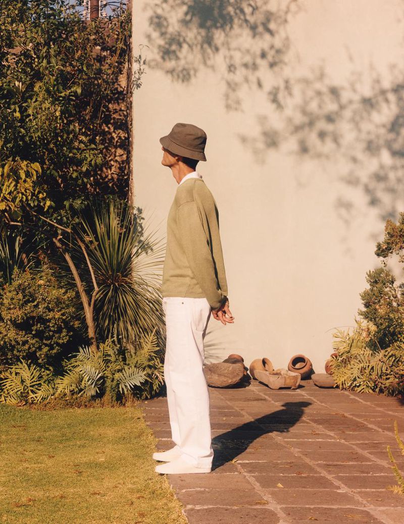 Side profile of a person in a green hat and sweater standing outdoors on a brick path with greenery and a white wall behind them.