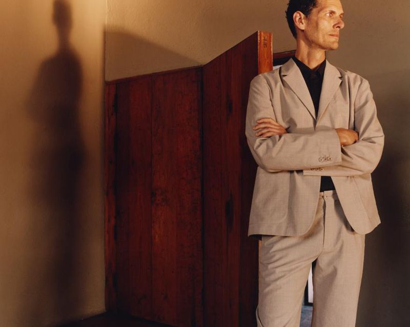Man in a suit stands indoors with arms crossed, beside a wood partition. A shadow is cast on the wall in a well-lit interior space.