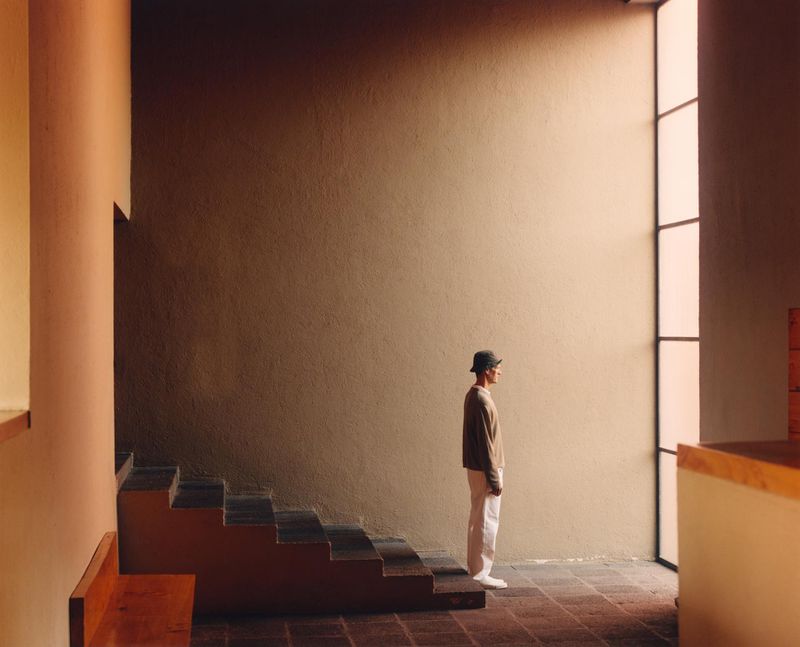 Man in tan shirt and white pants stands in minimalist building with light entering from a side window, casting shadows on staircase.