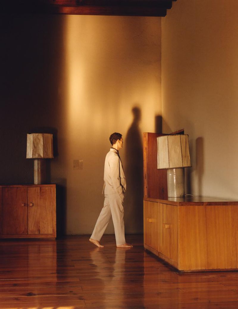 Man walking barefoot indoors past wooden cabinets and lamps in a warm-lit room for Zara Origins S/S 25