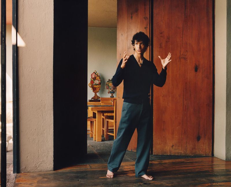 Barefoot man in V-neck sweater and blue pants poses indoors with decorative vases and wood accents in natural light, standing on wood floor.