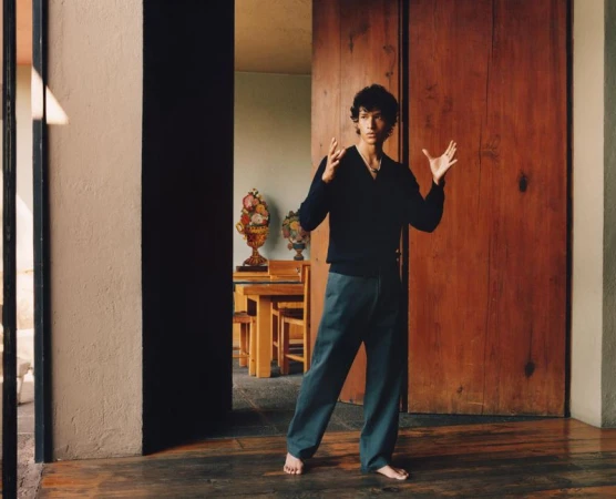 Barefoot man in V-neck sweater and blue pants poses indoors with decorative vases and wood accents in natural light, standing on wood floor.