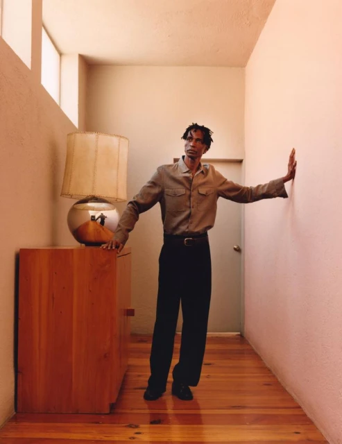 Man in gray shirt and black pants posing in hallway with lamp, wood floor, and peach wall. Light streams from window.