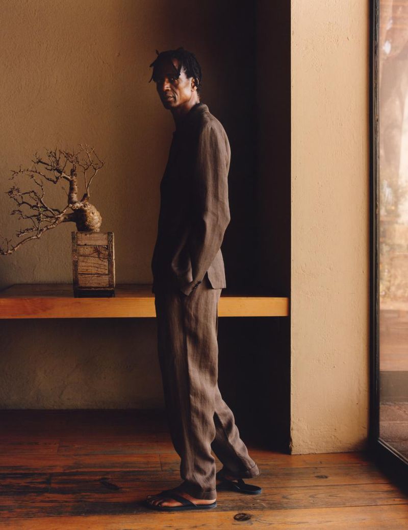 Dark-skinned adult male in brown suit poses in indoor setting with bonsai tree on wooden table, captured with natural light and eye-level angle.