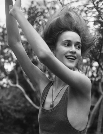 Black and white portrait of a smiling young woman with her arms raised, wearing a tank top and pendant necklace in an outdoor setting.