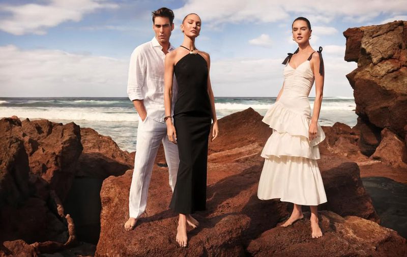 Three models pose on a rocky coast: a man in white linen, two women in black and white dresses, against a seascape backdrop.