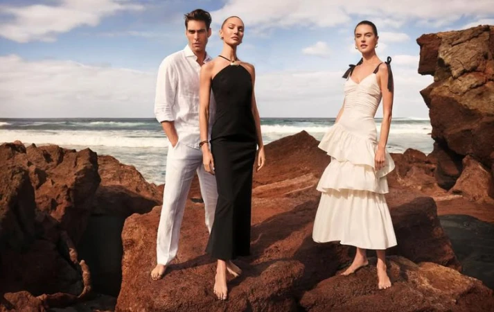 Three models pose on a rocky coast: a man in white linen, two women in black and white dresses, against a seascape backdrop.