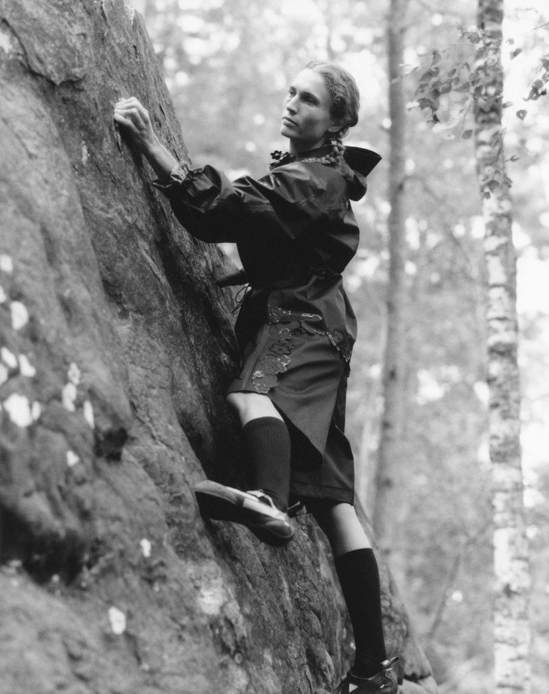 Monochrome photograph of a woman rock climbing on a textured stone surface in a wooded area, wearing outdoor attire and climbing shoes.
