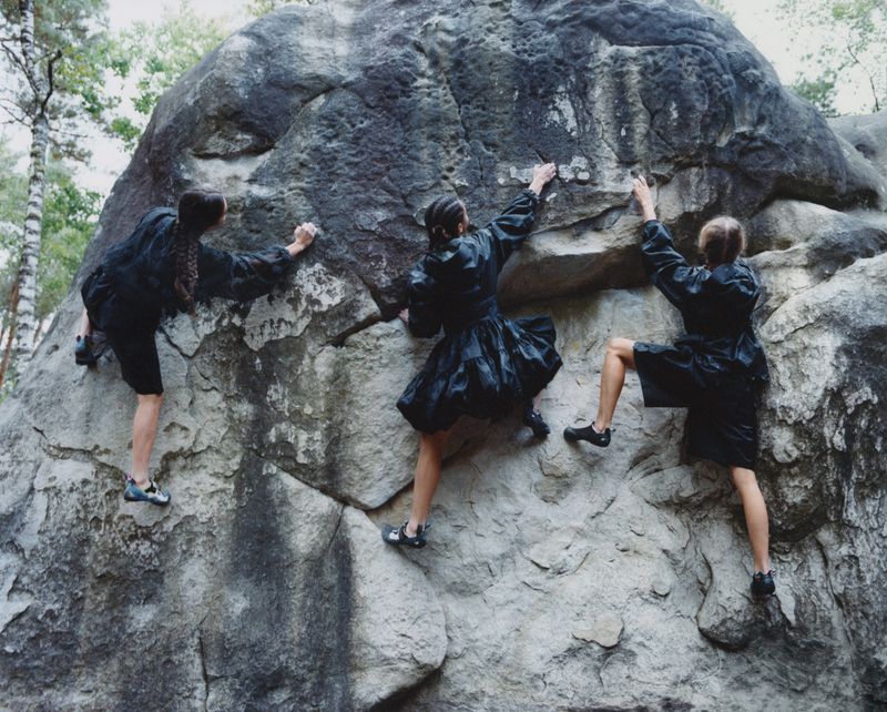 Three women in dark dresses rock climbing a large boulder outdoors surrounded by trees, captured in an eye-level medium shot.