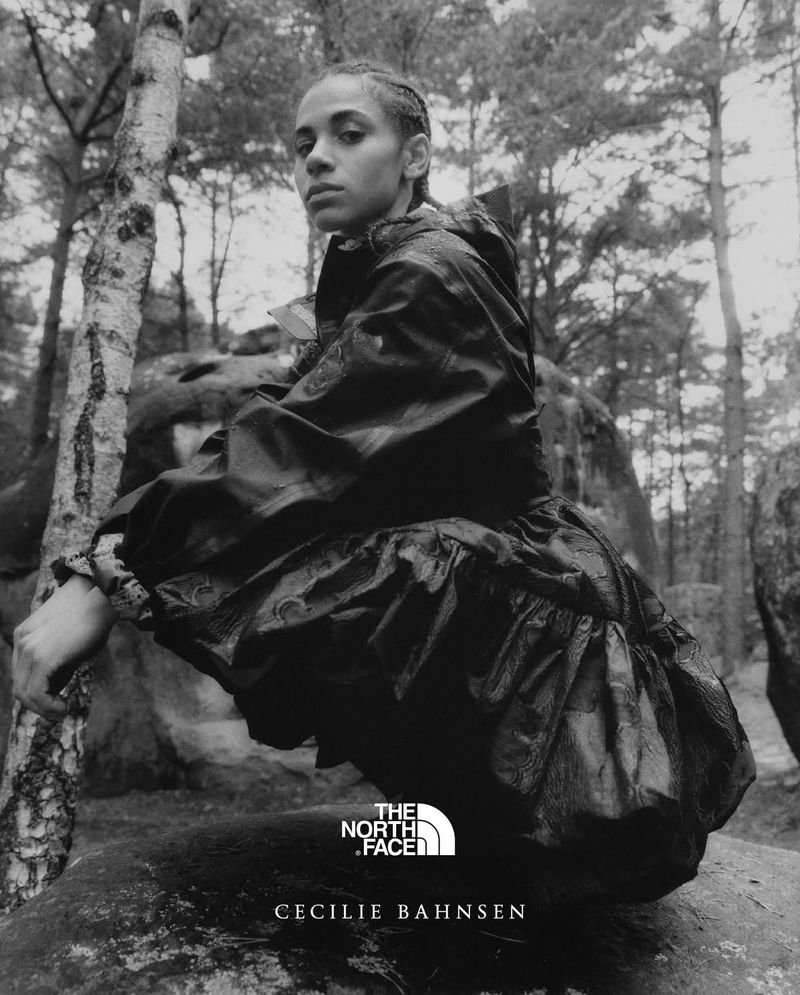 A woman in a black dress and jacket crouching on a rock in the forest. Trees and rocks surround her in this moody, grayscale fashion editorial.