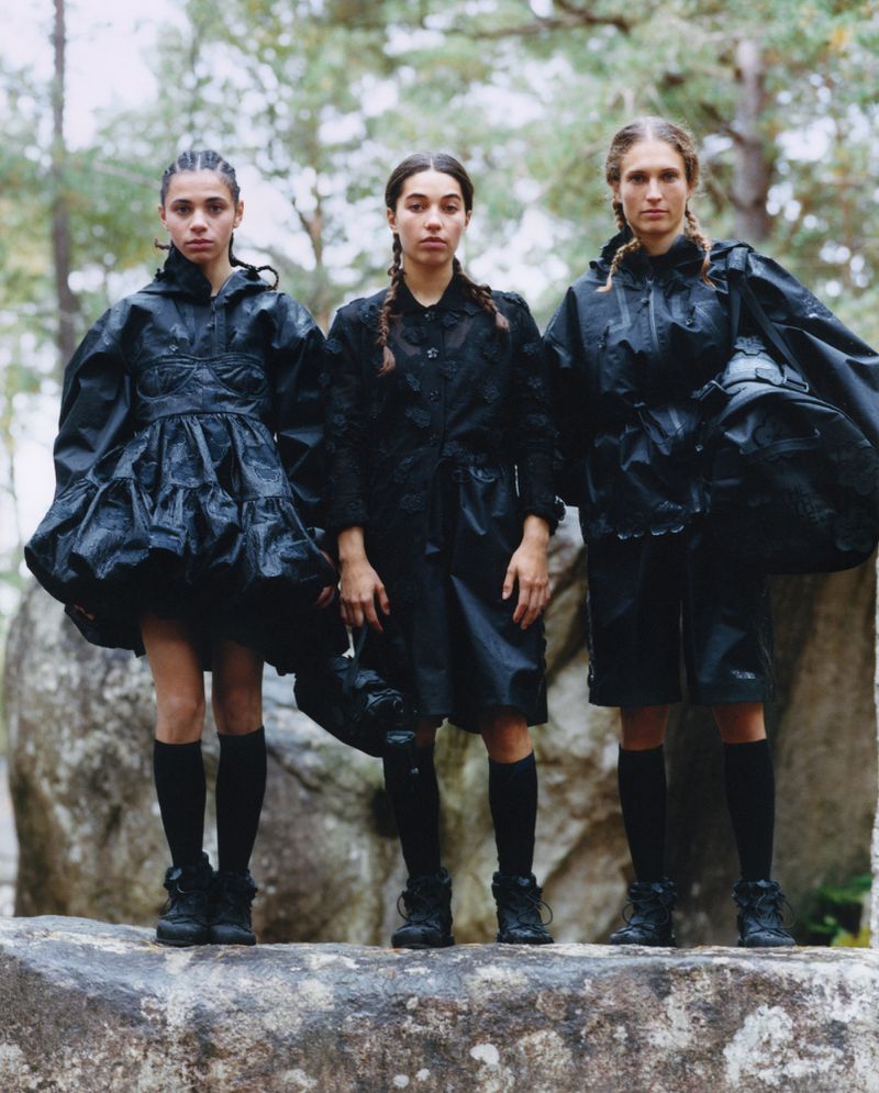 Three fashion models in matching black outfits pose on a rock in a wooded area, with trees in the background, showcasing dark, edgy style.