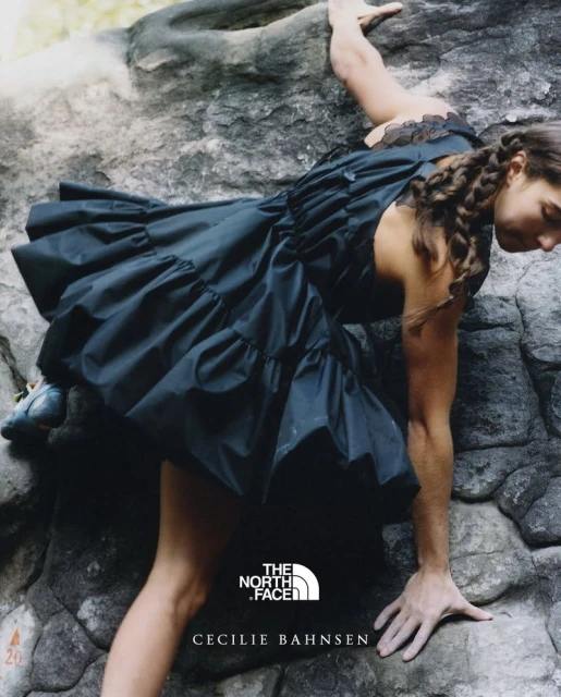 A woman in a black dress climbs a rock face in an outdoor setting, showcasing a blend of fashion and athleticism, with visible brand logos.