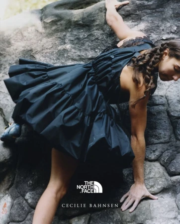 A woman in a black dress climbs a rock face in an outdoor setting, showcasing a blend of fashion and athleticism, with visible brand logos.