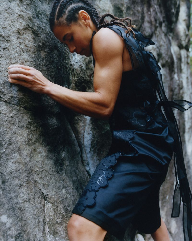 A woman in dark, fashionable clothing climbs a rock face outdoors, captured in a portrait-style shot with natural light and outdoor setting.