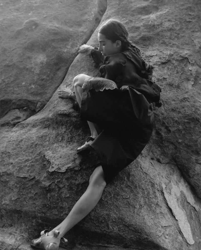 Young woman in a dark dress climbing a large rock formation in a black and white photograph, showcasing her strength and determination.